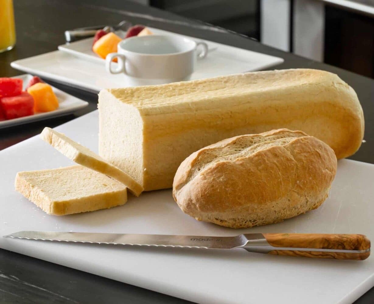 a loaf of white sandwich bread, sliced on a cutting board with a knife plus a small boule