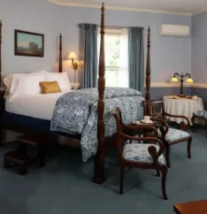 four poster bed with 2 chairs blue linens and 2 windows in bedroom