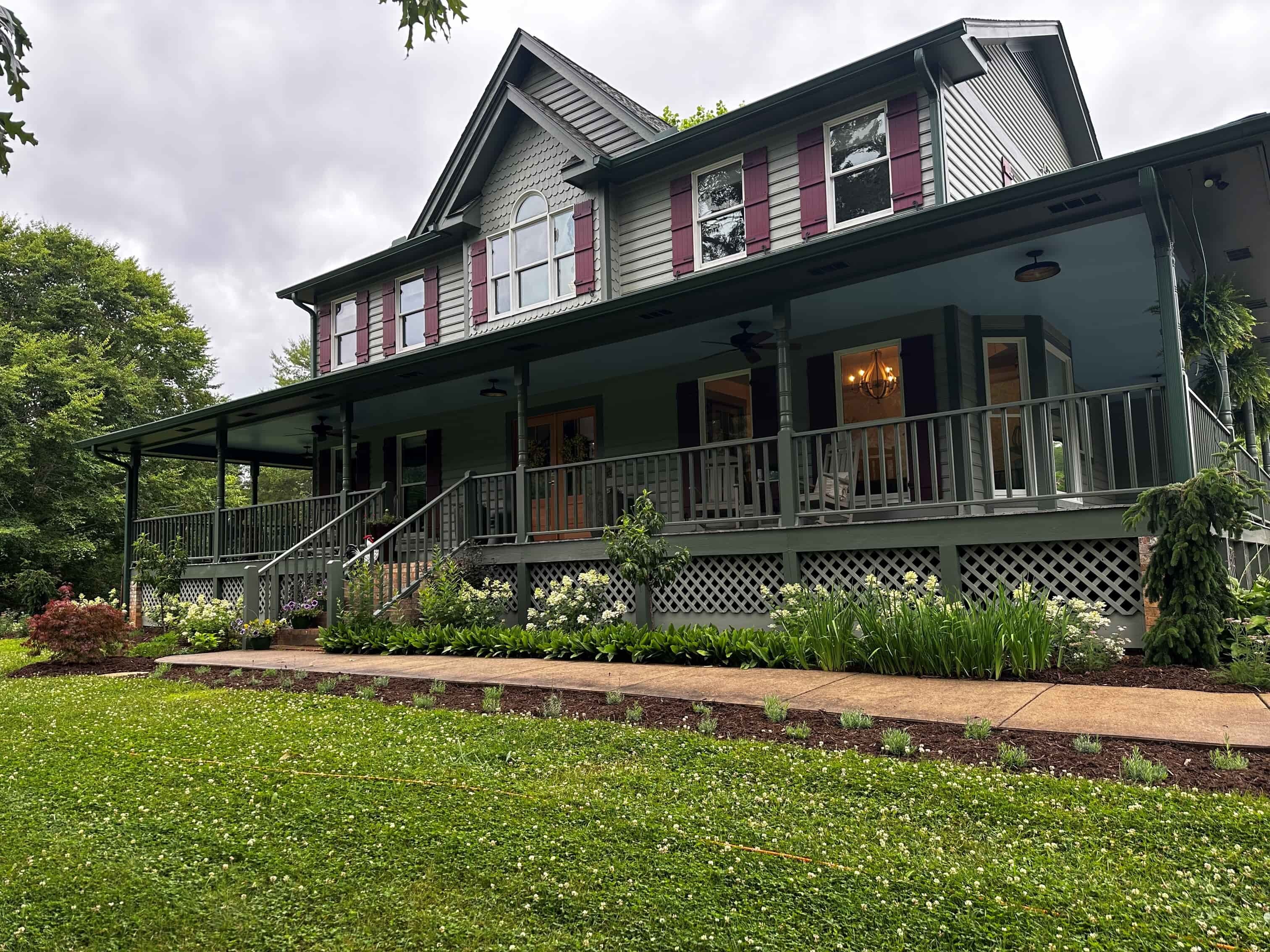 a gray 2-story farmhouse with burgundy shutters, wrap around porch, and sidewalk to greet guests at a B&B